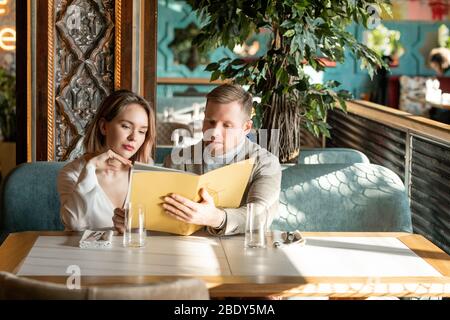 Young romantic couple looking through menu in luxurious restaurant and discussing what to choose while sitting on couch by table Stock Photo