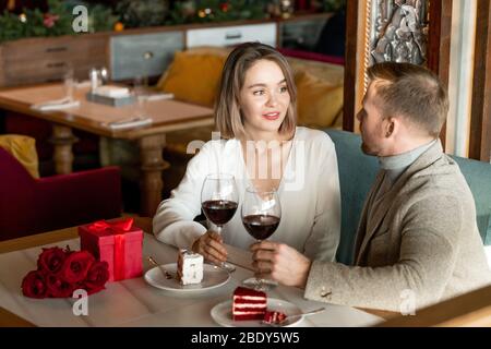 Young affectionate couple having talk by dessert and glass of red wine while sitting by served table after romantic lunch in restaurant Stock Photo