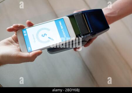 Hand of young man with smartphone paying by online banking while holding his mobile gadget over payment machine held by waiter Stock Photo