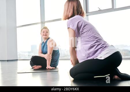 Happy disable girl in activewear looking at her friend while both sitting on mats and discussing exercises that they are going to do Stock Photo