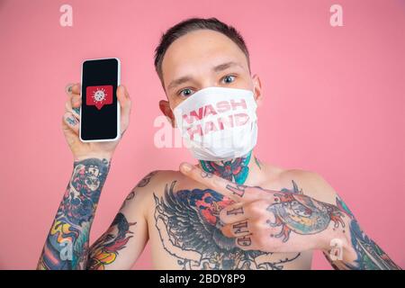 Young man with smartphone on pink background wearing face mask with words WASH HANDS. Concept of coronavirus spreading, protection, prevention, global epidemic and pandemic. Quarantine, isolation.