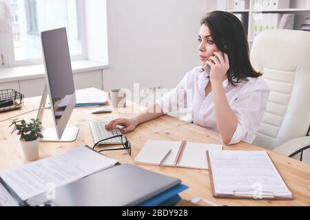 Busy young secretary of business company consulting clients on the phone while doing her work in front of computer monitor in office Stock Photo