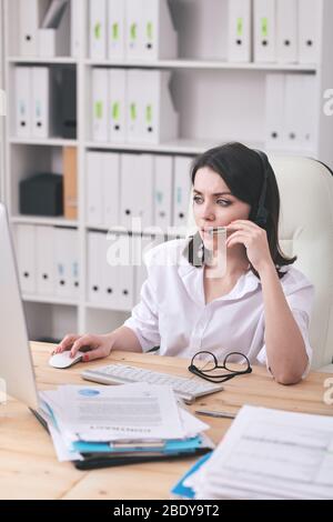 Pretty young serious operator with headset talking to clients and answering their questions online while working in front of computer Stock Photo
