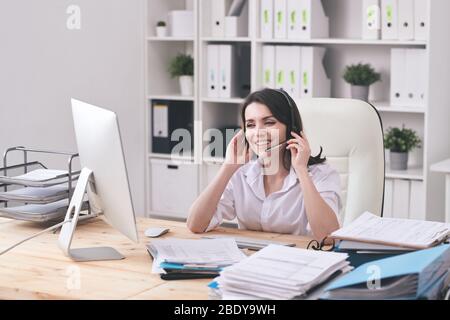 Happy young customer support representative with headset looking at client on computer screen and answering questions Stock Photo