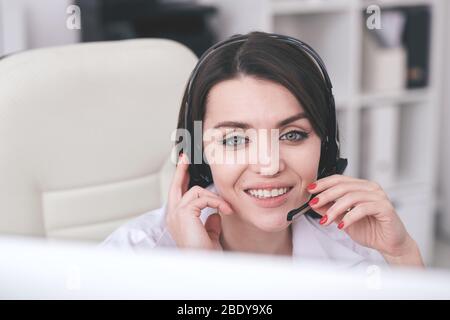 Young friendly telephone opeorator with toothy smile sitting in front of computer monitor and answering questions of clients online Stock Photo