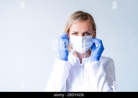 Blonde female doctor surgeon in white uniform, protective blue gloves putting on medical mask over grey. Healthcare, stay home. copyspace Stock Photo