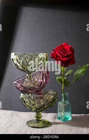 Three vases for cream of dark green and purple glass stand on top of each other on the black background with shadow. Dark red rose in the bottle inste Stock Photo