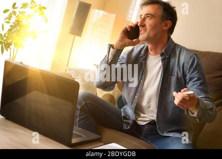 Freelance man working in the living room at home sitting on the sofa talking on mobile phone in a warm environment gesturing. Stock Photo