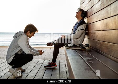 Two young friends with headphones and a skateboard laugh sitting bay the beach Stock Photo