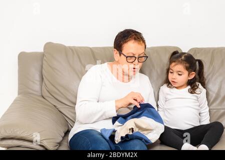 Grandmother Teaches Her Granddaughter To Mend Clothes. Kid Being Homeschooled Due To School Shut Down During Covid-19 Pandemic. Stock Photo