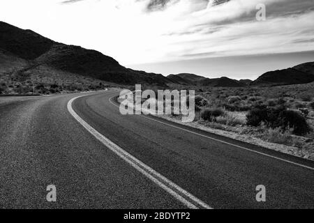 road with double yellow stripe vanishes in distance through the desert landscape. Mountains on the horizon and dotted clouds overhead. Stock Photo