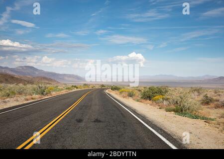 road with double yellow stripe vanishes in distance through the desert landscape. Mountains on the horizon and dotted clouds overhead. Stock Photo