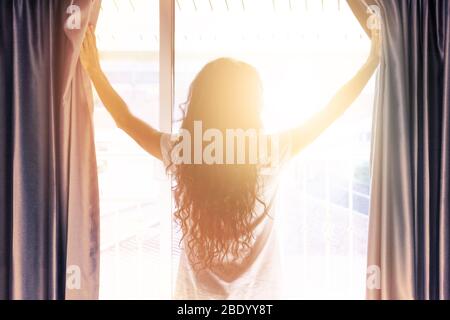 Woman opens the curtains in the morning in the bedroom.  Sunlight shines into the room. Concept of new day beginning. Blurred photo with soft focus. Stock Photo