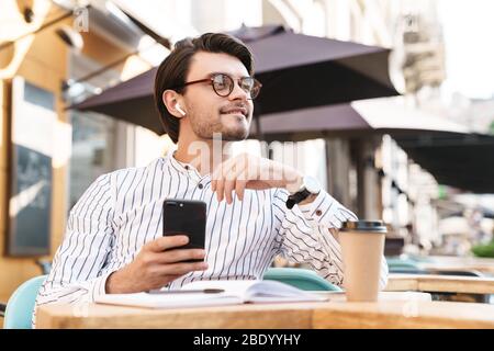 Photo of smiling caucasian man wearing eyeglasses using earpod and cellphone while working in cafe outdoors Stock Photo