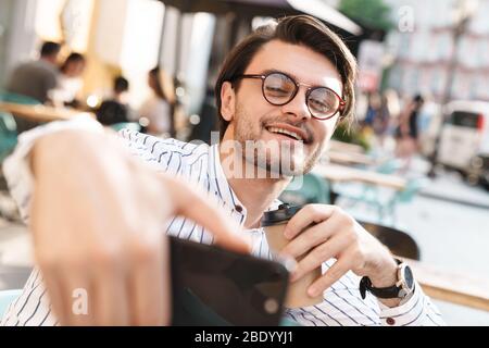 Photo of happy young man wearing eyeglasses drinking coffee and taking selfie photo on cellphone while working in cafe outdoors Stock Photo