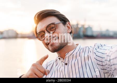 Photo of successful smiling man wearing eyeglasses pointing finger at camera and taking selfie photo while walking on promenade Stock Photo