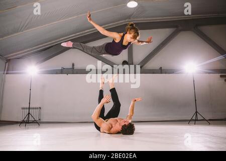 young couple Couple practicing acro yoga in gym. Healthy lifestyle. Acroyoga. Couple yoga. Partner yoga. Muscular man throwing women to air. Concept Stock Photo