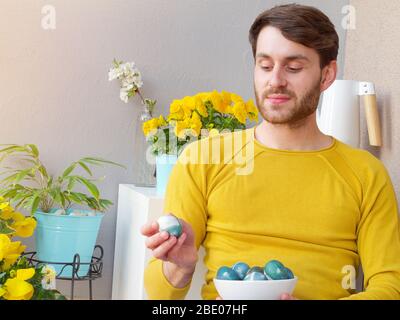 Caucasian man holding a blue easter eggs in a bowl during easter time, colored with natural red cabbage coloring, wearing a yellow sweather. Stock Photo