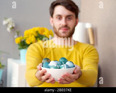 Caucasian man holding a blue easter eggs in a bowl during easter time, colored with natural red cabbage coloring, wearing a yellow sweather. Stock Photo