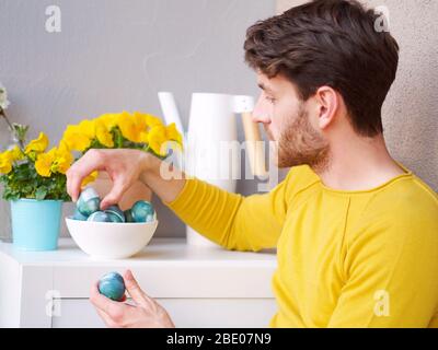 Caucasian man holding a blue easter eggs in a bowl during easter time, colored with natural red cabbage coloring, wearing a yellow sweather. Stock Photo