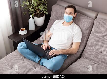 Happy relaxed young caucasian man working from home wearing protective mask using laptop. Cozy Home office, workplace on sofa during coronavirus Stock Photo
