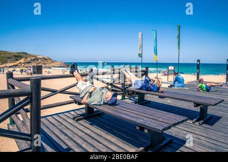 Two people resting with their legs up lying down on benches while others play at the beach at Praia Das Macas Portugal Stock Photo
