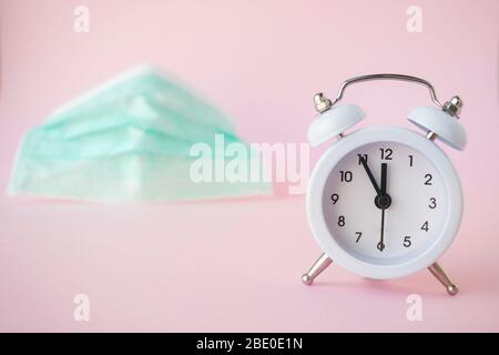 Time to buy medical/surgical concept. Small white clock showing five minutes to 12, with face mask on the light pink background. Outbreak coronavirus. Stock Photo