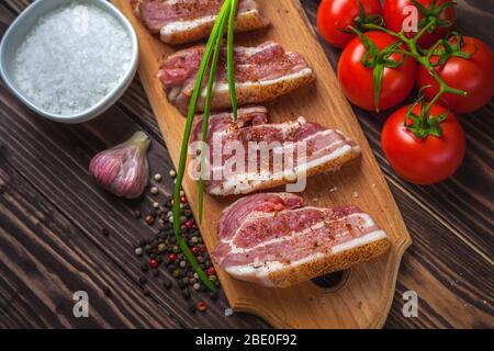 Raw slices of pork bacon on a cutting board, on a rural wooden tabletop. Pork belly with vegetables. Stock Photo