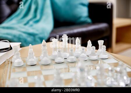 Close up of frosted glass chess pieces on a glass chess board in a living room with reading glasses and a hardcover book and shallow depth of field Stock Photo