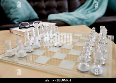 Close up of frosted glass chess pieces on a glass chess board in a living room with reading glasses and a hardcover book and shallow depth of field Stock Photo