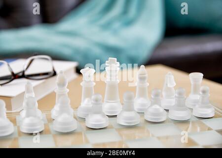 Close up of frosted glass chess pieces on a glass chess board in a living room with reading glasses and a hardcover book and shallow depth of field Stock Photo