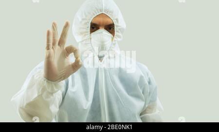 Front view of doctor with mask and bioprotective suit with arm extended forward and his right hand making an OK sign on white background Stock Photo
