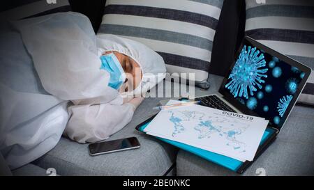Close up view of doctor with blue mask and bioprotective suit sleeping next to his smart phone, a laptop with the image of coronavirus on the screen a Stock Photo