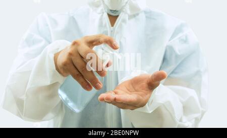 Doctor with mask and white bioprotective suit washing his hands with a bottle of alcohol gel in the foreground on white background Stock Photo