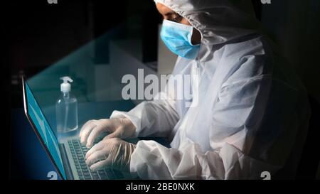 Side view of doctor with blue face mask and concentrated bioprotective suit working in office with his laptop on his desk with bottle of alcohol gel w Stock Photo