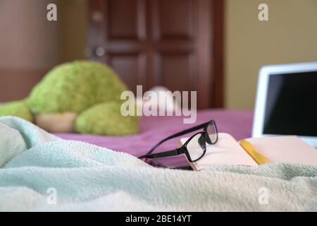 Computer laptop with notepad, pen, reading glasses and soft toy on top of bed in the bedroom. Work at home. Focus on spectacles. Stock Photo