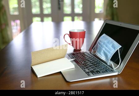 Work at home during coronavirus pandemic. Focus on face mask on computer laptop together with notepad and red coffee cup on the dining table. Windows Stock Photo
