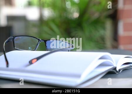 Reading glasses on top of an open book. Garden or green nature background. Stock Photo
