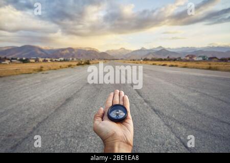 Traveler holding compass on the road with mountains at sunrise sky background. Travel and adventure concept. Stock Photo