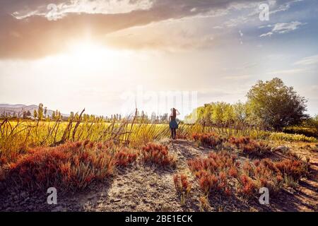 Woman in hat and green checked shirt near the fence looking at field in the farm at sunset cloudy sky background
