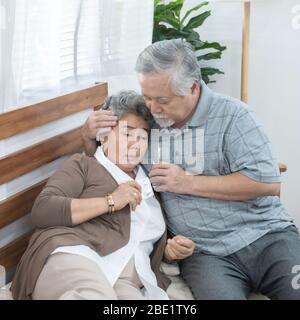 Asian Senior Woman taking medicines and drinking water while sitting on couch. Old man take care his wife while her illness at the house. Stock Photo