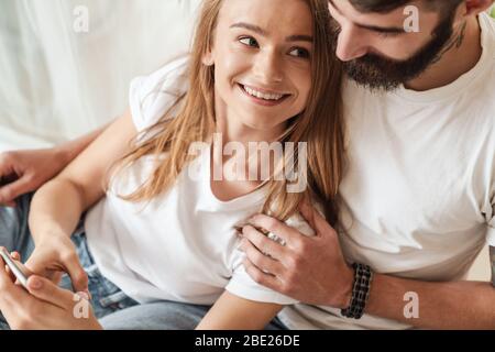 Image of happy young couple smiling and using cellphone while hugging in bright room at home Stock Photo