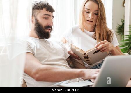 Image of caucasian serious couple using laptop and reading book while sitting on couch in bright room at home Stock Photo
