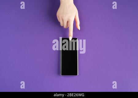 Beautiful female hand with white manicure pointing on contemporary smartphone of black color on purple background. Flat lay. Place for text. Mockup fo Stock Photo