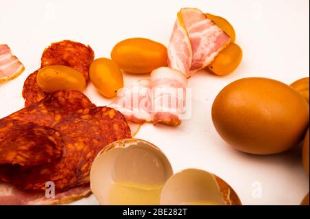 Broken chicken egg, scattered chicken eggs, slices of sausage and bacon and tomatoes on a white background. Close up Stock Photo