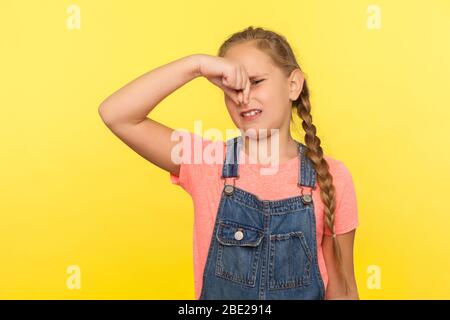 Awful smell. Portrait of little girl with braid in denim overalls holding breath, pinching her nose and expressing disgust to stink, intolerable odor. Stock Photo