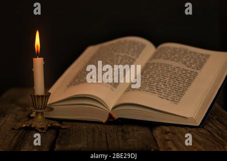 Open Torah and a burning candle in a candlestick on a wooden surface on a dark background close up Stock Photo