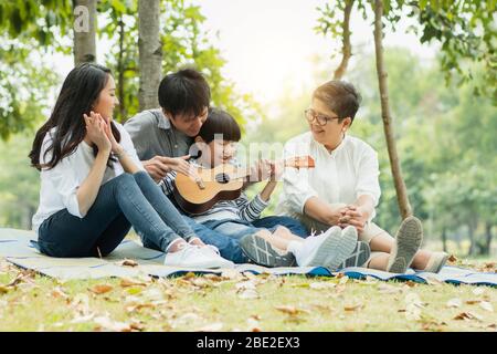 Happy family with grandma, mom with dad teaching son  playing guitar and sing a song in park, Enjoy and relax people picnic outside Stock Photo