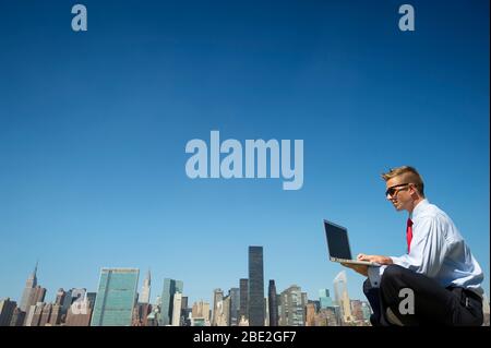 Businessman in a suit sitting outdoors using laptop computer above the city skyline under clear blue sky copy space Stock Photo