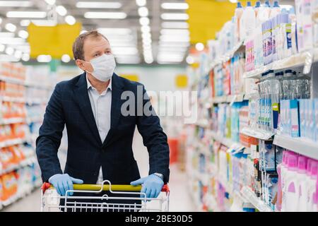 Serious attentive man chooses household detergents in store, walks with shopping cart, buys necessary products during quarantine, wears protective med Stock Photo
